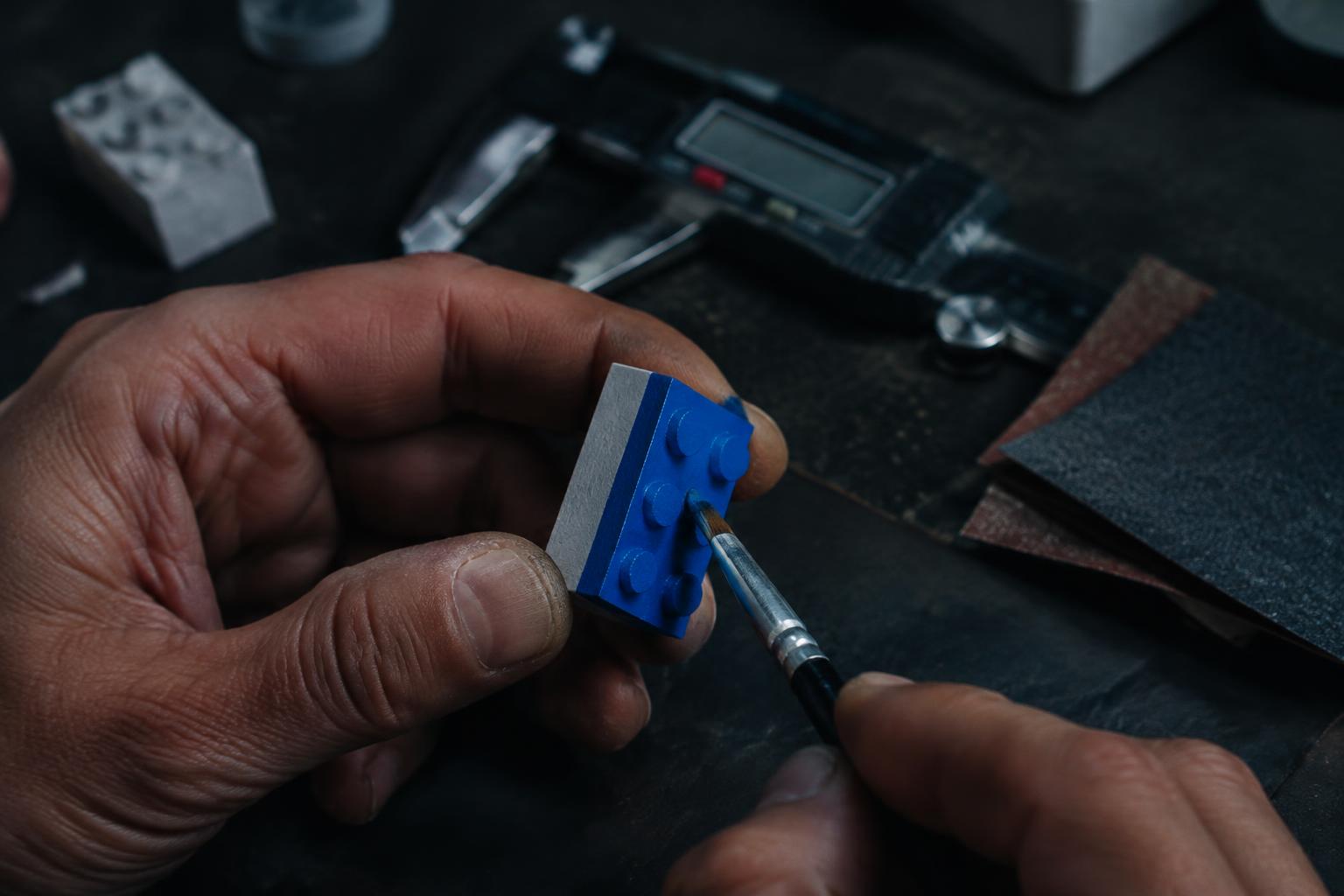 Hands sanding and painting a freshly 3D printed LEGO-compatible brick beside digital calipers