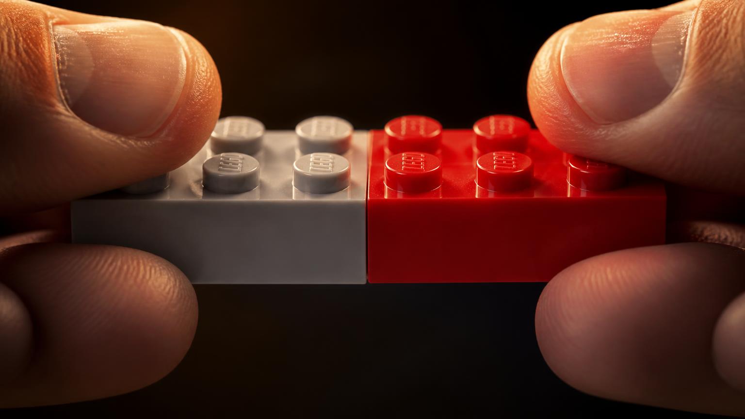 Macro close-up of fingers pressing a grey and a red 2x4 compatible brick together to test clutch power and stud tolerance